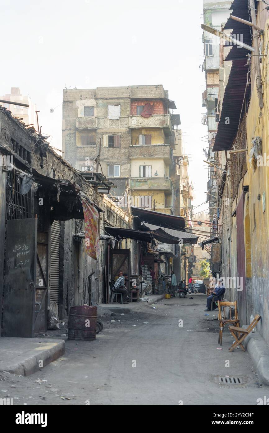 Enge Gasse in Alexandria, Ägypten, gesäumt von alten Gebäuden, Straßenläden und Bewohnern, die sich auf Stühlen entspannen und das urbane ägyptische Leben einfangen. Stockfoto