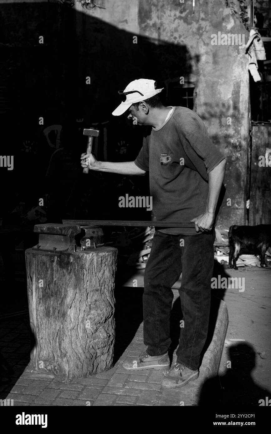 Schmied arbeitet in Alexandria, Ägypten, hämmert Metall in einer rustikalen Werkstatt. Ein markanter Kontrast von Licht und Schatten unterstreicht seinen Fokus und sein Geschick. Stockfoto