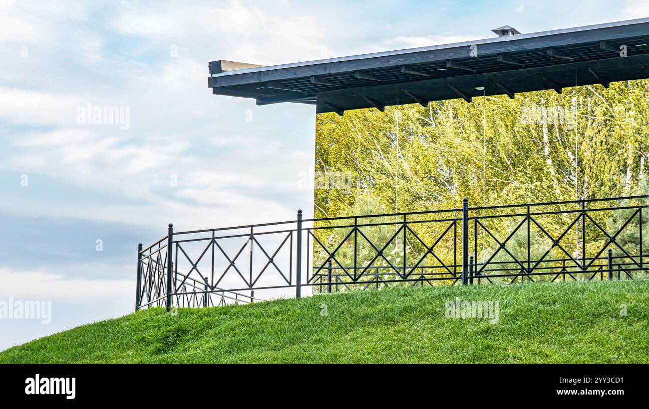 Das moderne Spiegelgebäude spiegelt Bäume auf einem grünen Hügel mit einer schwarzen Schiene wider. Unter blauem, bewölktem Himmel verbindet Architektur mit Natur Stockfoto