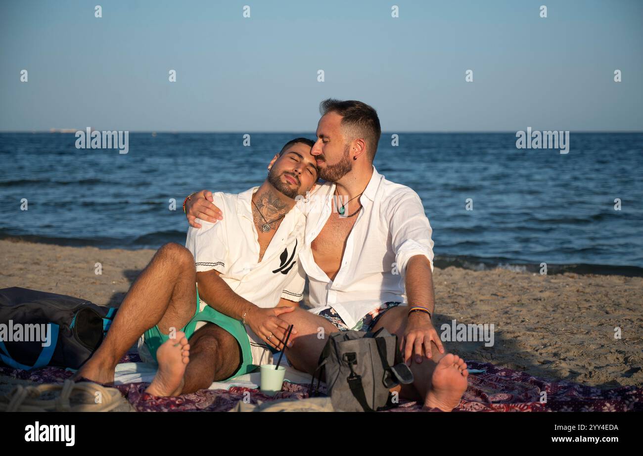 Ein junges schwules Paar genießt einen ruhigen Moment am Strand und zeigt Liebe und Intimität. Es ist entspannt auf einer Decke und genießt die Schönheit des küstenraumes Stockfoto