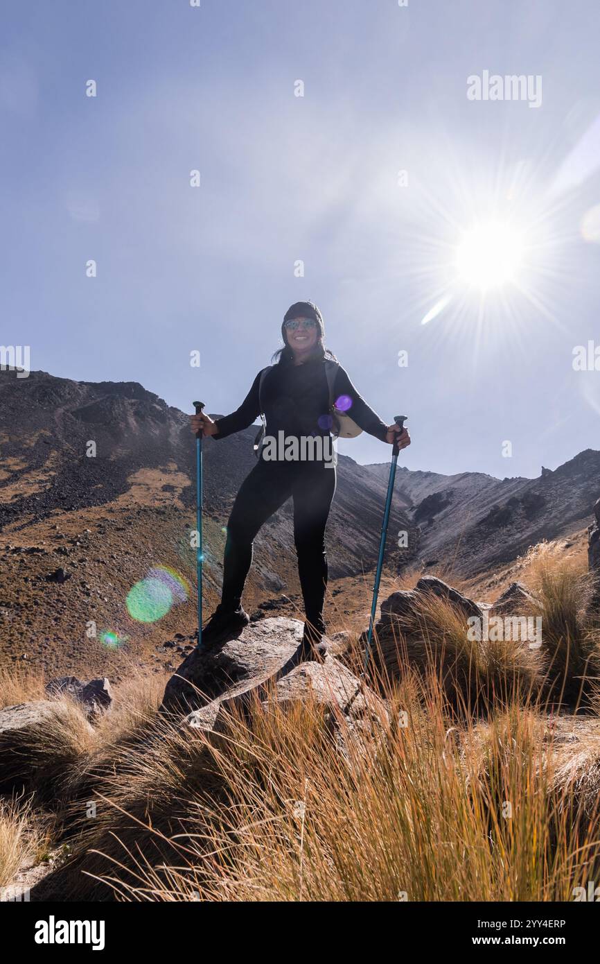 Ein Wanderer steht selbstbewusst auf einem Felsvorsprung und hält Wanderstöcke. Die Sonne scheint hell und wirft Leuchtgläser, die den zerklüfteten Berg beleuchten Stockfoto