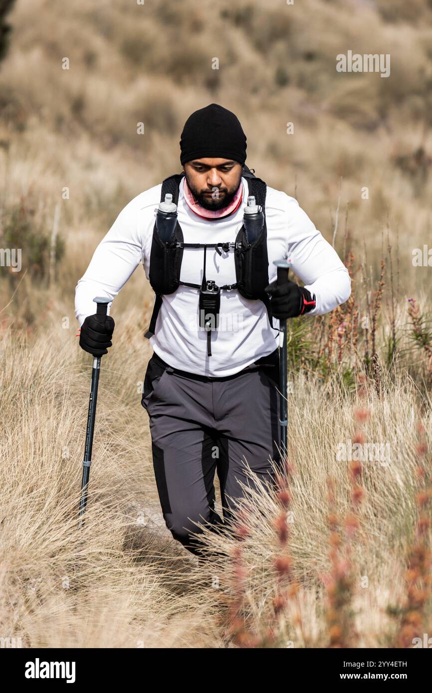 Ein schwarzer Mann mit fortgeschrittener Bergsteigerausrüstung, einschließlich einer weißen Jacke und einem Rucksack, wandert durch eine grasbewachsene Landschaft. Er hält Trekkingstöcke, foc Stockfoto
