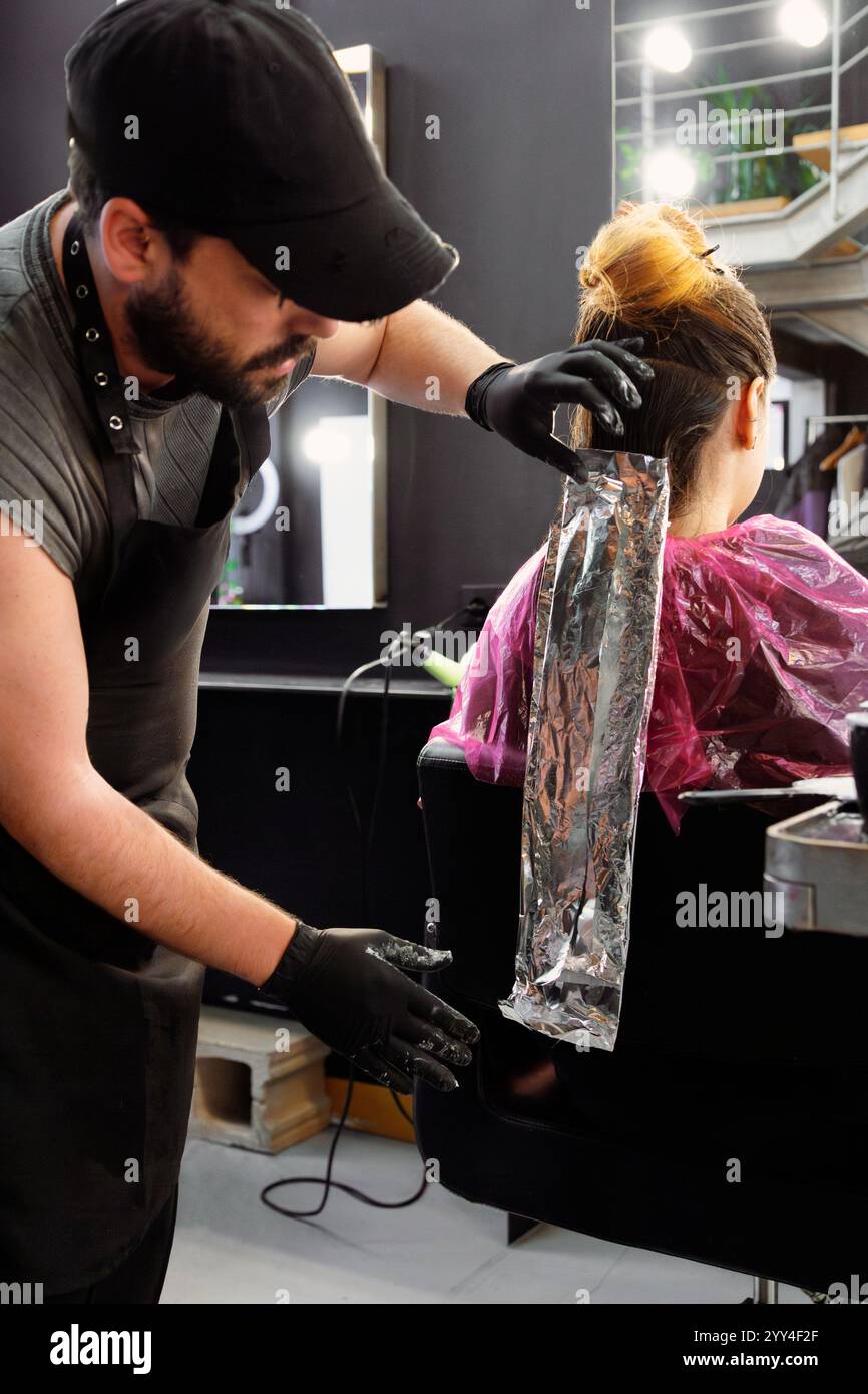 Ein Friseur mit Handschuhen trägt die Haarfarbe mit Folien in einem modernen Salon. Der Kunde sitzt in einem Stuhl mit einem cape, umgeben von einem eleganten, stilvollen de Stockfoto