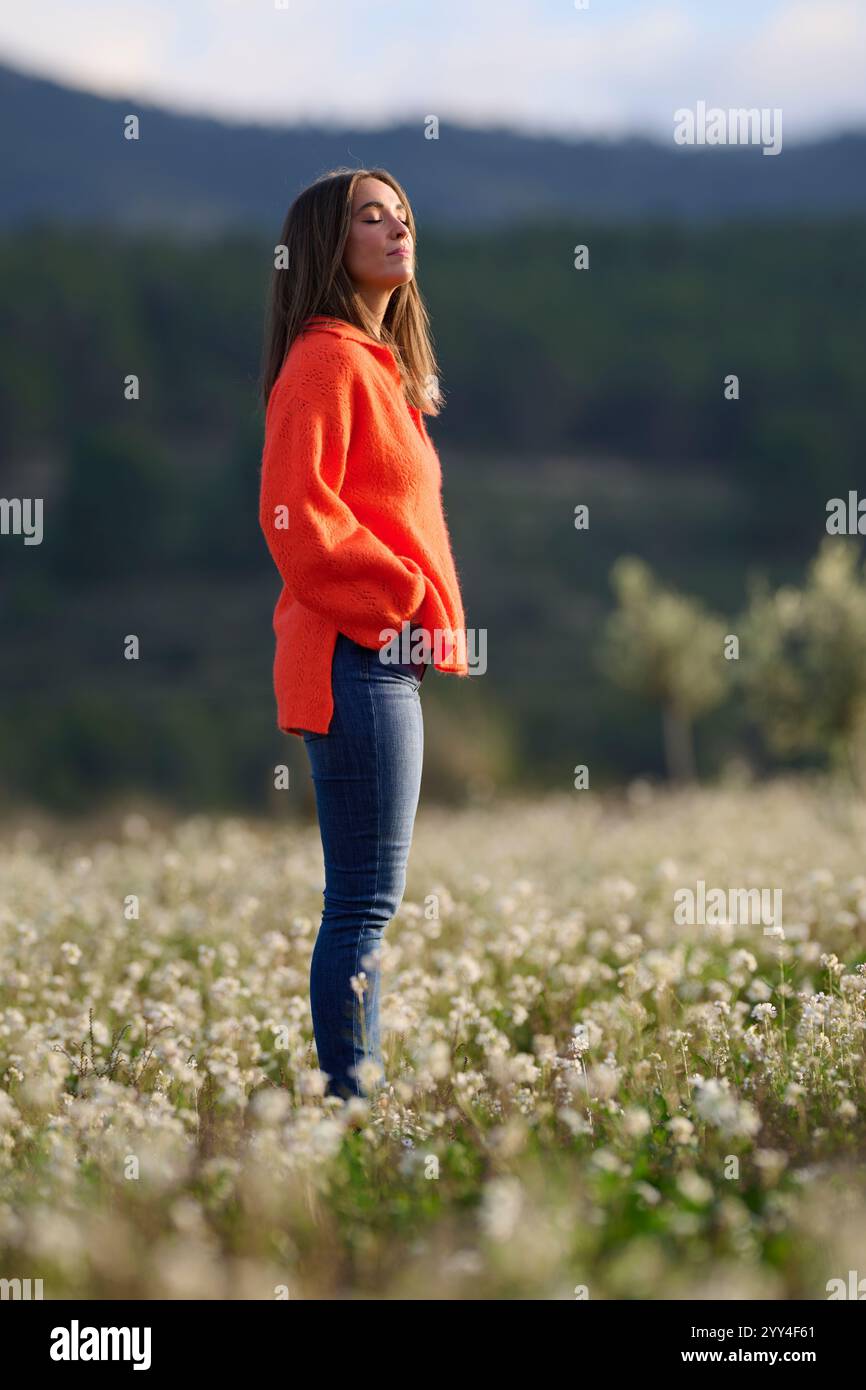 Eine Frau in einem leuchtend orangefarbenen Pullover steht auf einer sonnendurchfluteten Wiese, umgeben von Wildblumen. Sie wirkt nachdenklich und entspannt, mit sanft geschlossenen Augen Stockfoto