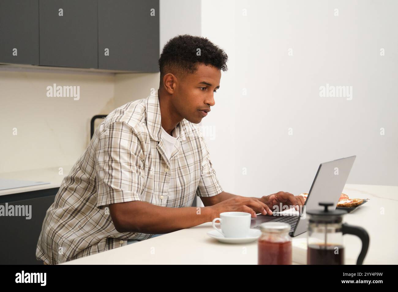 Ein junger afroamerikanischer Mann genießt ruhige Momente zu Hause. Der Mann konzentriert sich auf einen Laptop, während die ruhige, moderne Küche zur gemütlichen Atmosphäre beiträgt Stockfoto