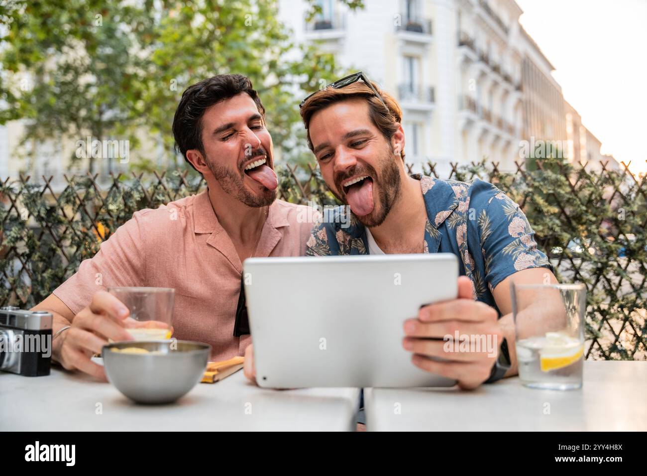 Ein fröhliches schwules Paar sitzt in einem Café im Freien, hält spielerisch die Zunge aus, während es ein digitales Tablet verwendet, um einen Moment des Spaßes und der Verbindung zu zeigen Stockfoto