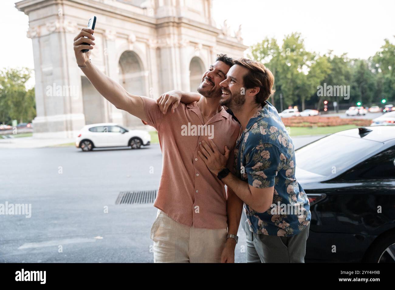 Ein fröhliches schwules Paar steht auf einer geschäftigen Stadtstraße und hält mit einem Smartphone einen liebevollen Moment in einem Selfie fest Stockfoto
