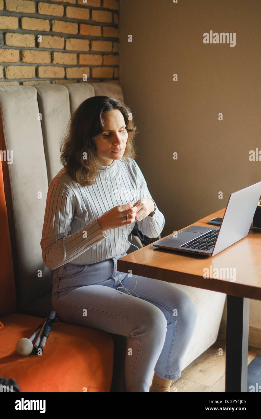 Eine blinde Frau sitzt an einem Holztisch und arbeitet in einem Café mit einem Laptop und Kopfhörern. Sie scheint konzentriert zu sein, mit einem weißen Stock an ihrer Seite, der Technik umschließt Stockfoto