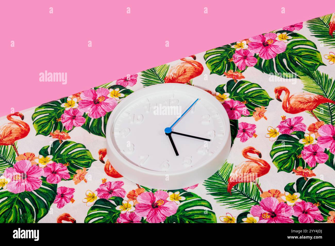 Eine minimalistische weiße Wanduhr liegt auf einem lebhaften tropischen Stoff mit Flamingos, Hibiskusblüten und Palmblättern. Der farbenfrohe Hintergrund-cre Stockfoto