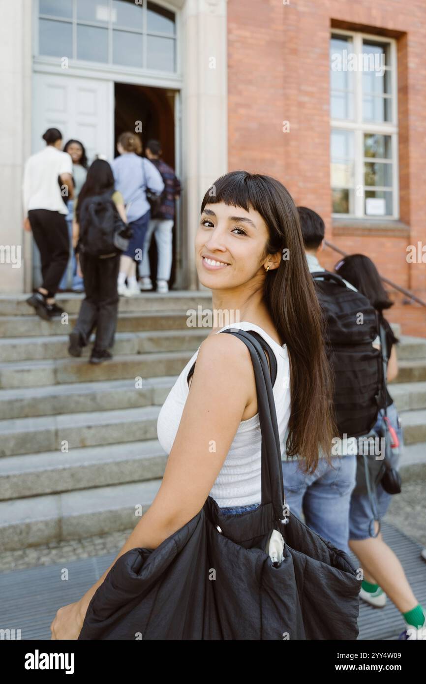 Porträt einer lächelnden Frau, die Tasche trägt, während sie auf dem College-Campus über die Schulter schaut Stockfoto