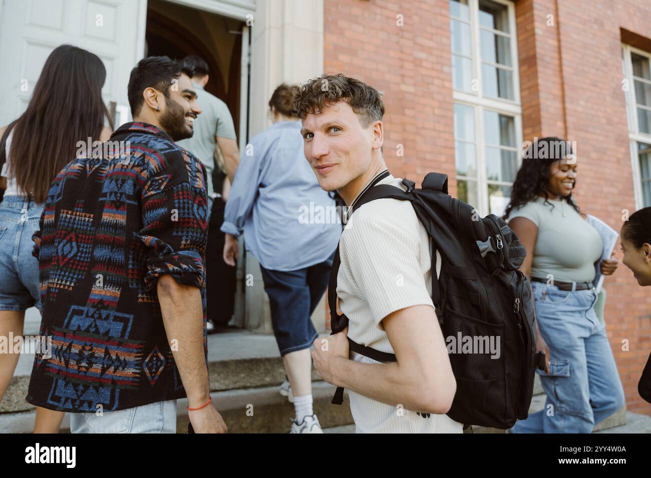 Porträt eines lächelnden Mannes, der seine Tasche trägt und die Treppe hinaufzieht, während er über die Schulter auf dem College-Campus schaut Stockfoto
