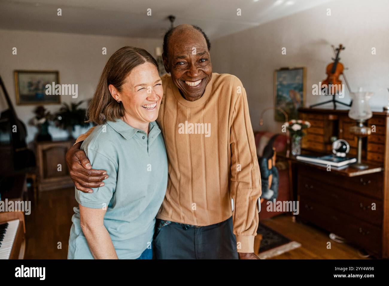 Porträt eines glücklichen älteren Mannes, der mit Arm um eine Krankenschwester im Wohnzimmer zu Hause steht Stockfoto