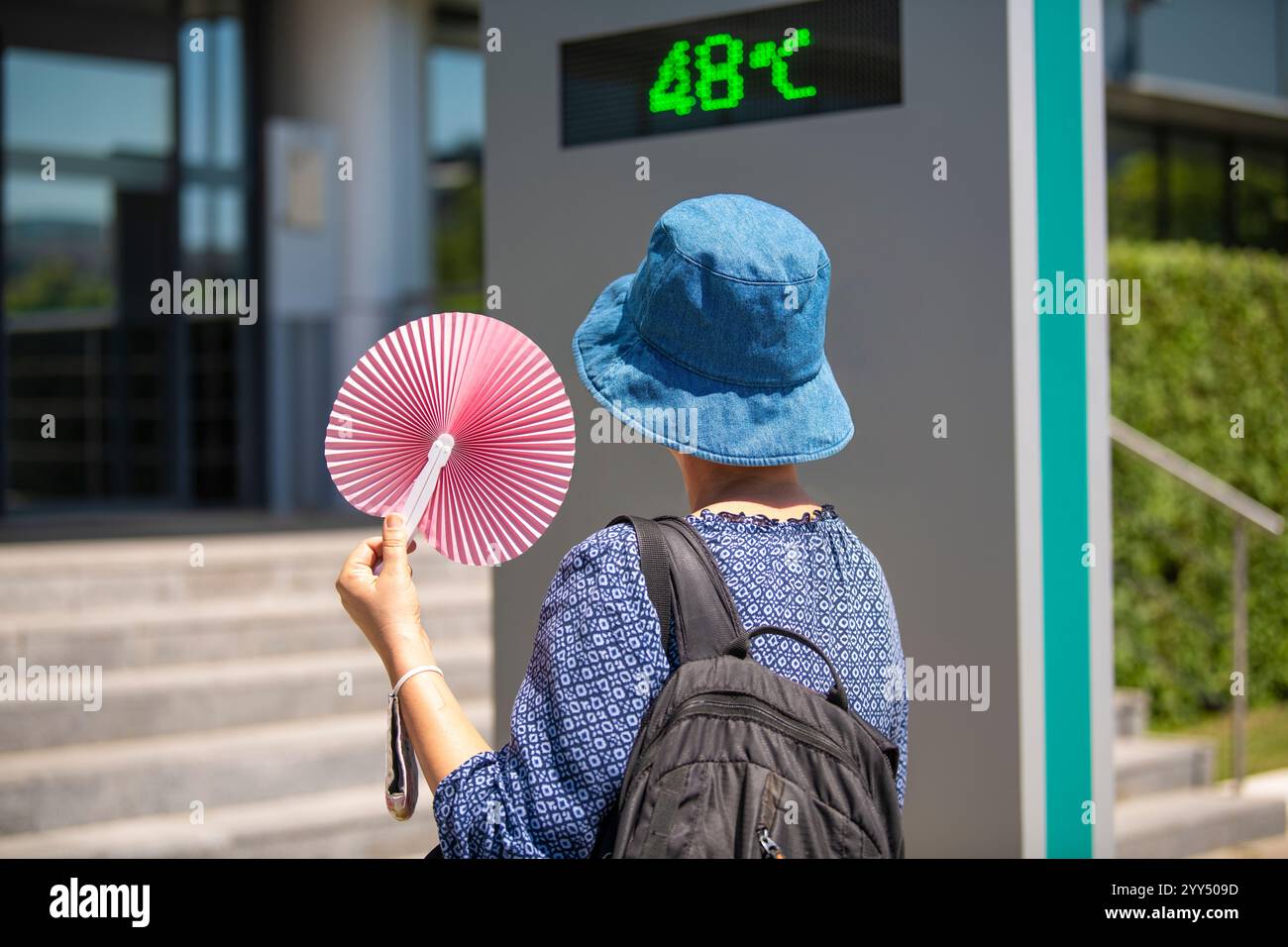Frau mit Hut und Papierwinkelventilator, die auf Straßenthermometer und extreme Temperaturmarkierung von 48 Grad celsius an heißen Sommertagen blickt. Stockfoto