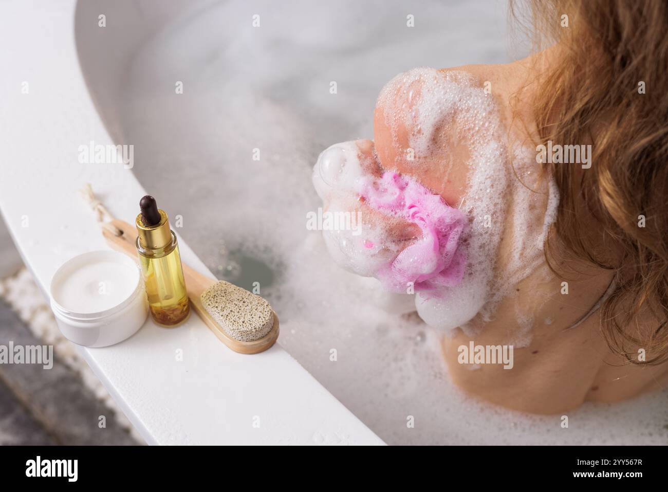 Glückliche junge Frau, die Duschgel mit Luffa-Schwamm auf ihren Körper aufträgt, während sie duscht. Spa- und Körperpflegekonzept, Stockfoto