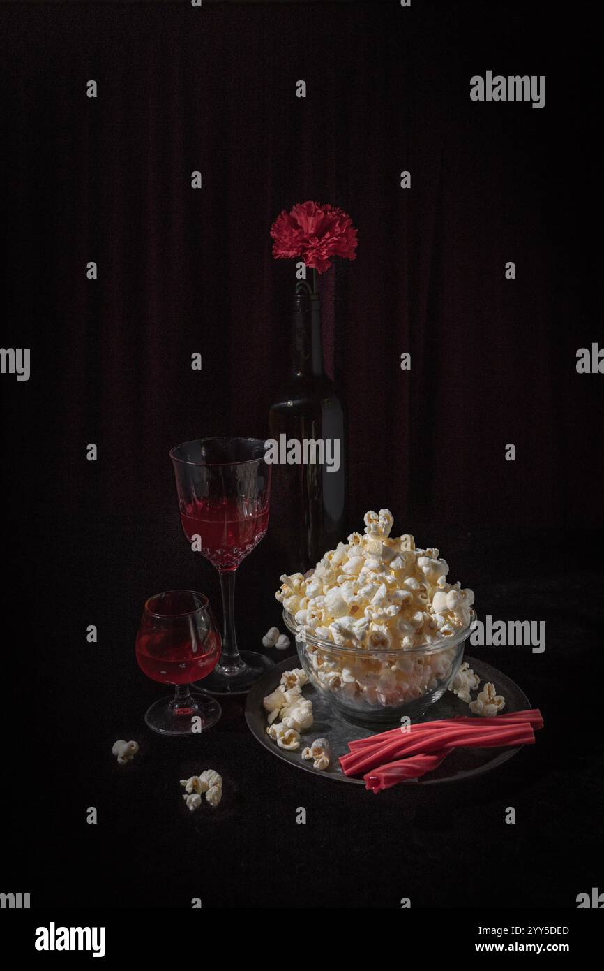 Ein vom Kino inspiriertes Stillleben mit einer Schüssel Popcorn, roten Getränken in eleganten Gläsern und einer Flasche mit roter Nelke auf der Oberseite. Das Setup ruft hervor Stockfoto