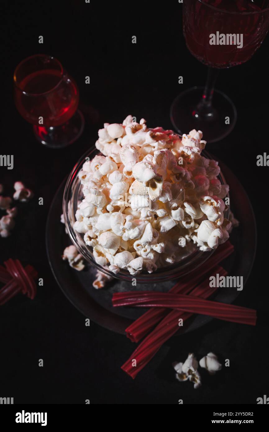 Ein flacher Blick auf Popcorn in einer Schüssel, rote Getränke in Gläsern und rote Lakritzstäbchen auf dunklem Hintergrund. Das Arrangement vermittelt einen gemütlichen Filmabend Stockfoto