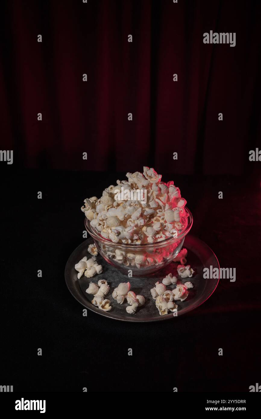 Eine einzelne Schüssel mit frisch gepufftem Popcorn vor dunklem Hintergrund mit dezenten Rottönen, die an einen Theatervorhang erinnern und an ein filmisches atmos erinnern Stockfoto