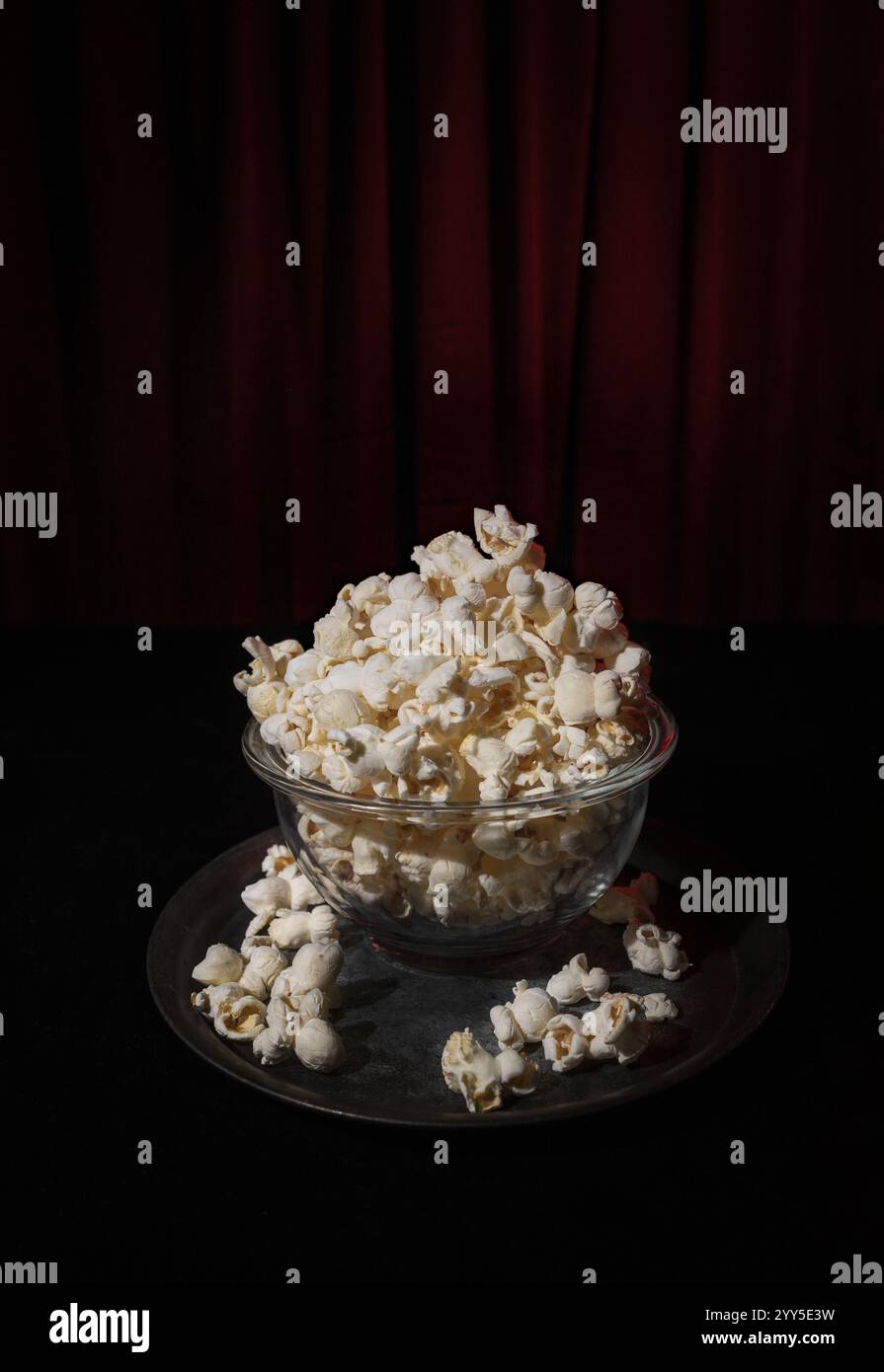 Eine einzelne Schüssel mit frisch gepufftem Popcorn vor dunklem Hintergrund mit dezenten Rottönen, die an einen Theatervorhang erinnern und an ein filmisches atmos erinnern Stockfoto