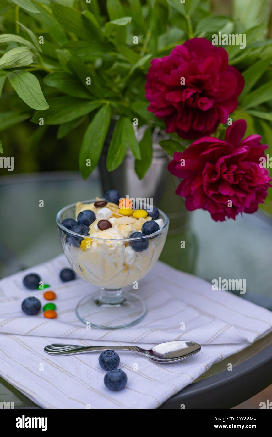Köstliches Vanilleeis in einer Glasschale mit frischen Blaubeeren, bunten Süßigkeiten und Mandelscheiben auf einer weißen Serviette, mit leuchtenden roten Pfingstrosenblüten Stockfoto