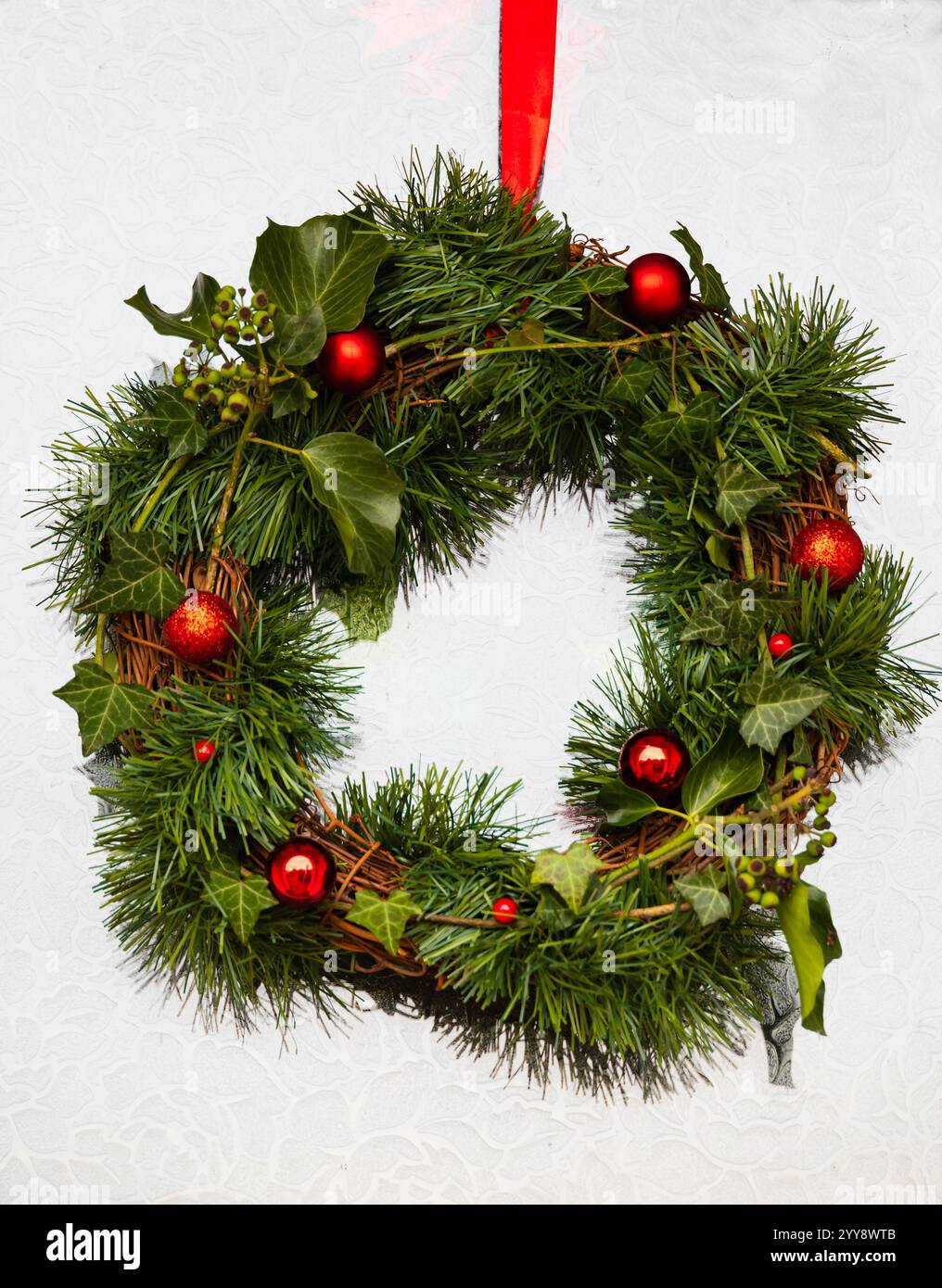 Hausgemachter, handgemachter natürlicher Weihnachtskranz auf mattiertem weißem Glas-Vordertür. Efeu, Tanne und rote Kugeln. Stockfoto