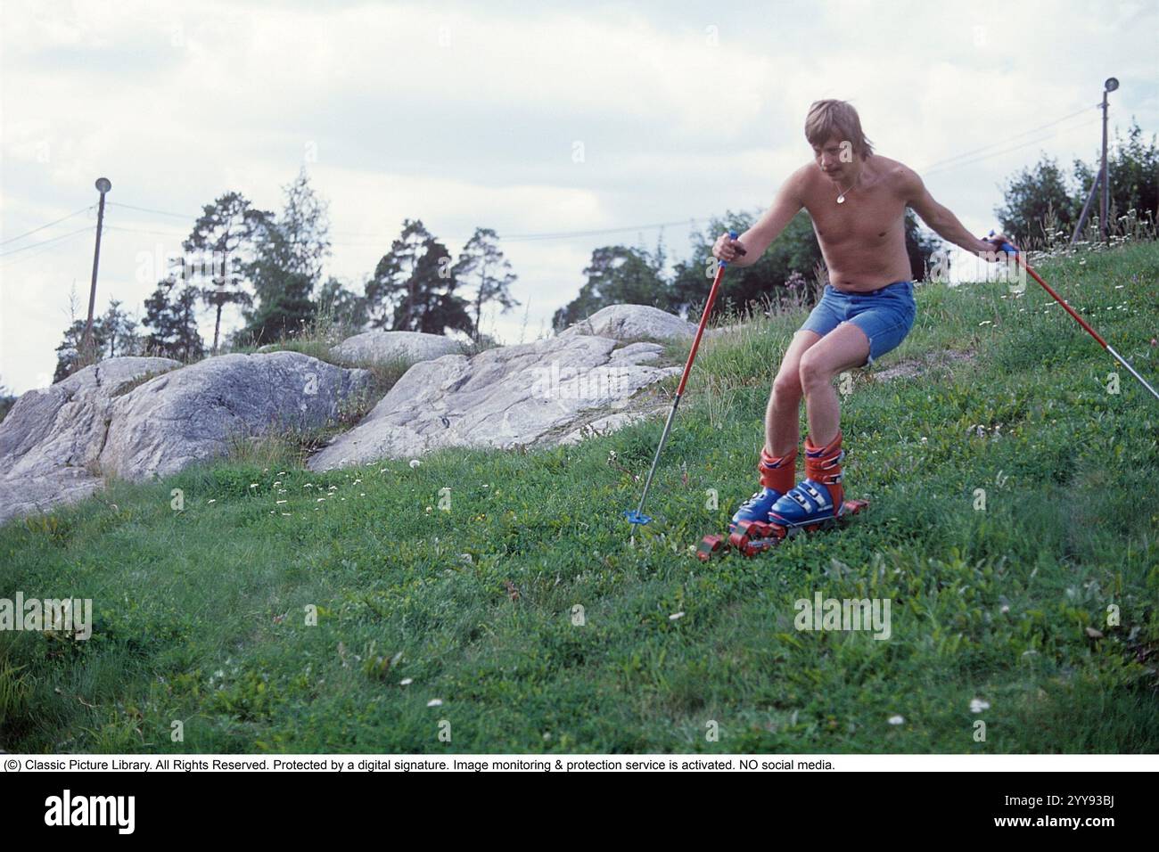 Slalom ohne Schnee. 1980 wird im Sommer eine neue Erfindung der Grasskier getestet. Die Skifahrer sind mit Rollbändern ausgestattet und hatten ähnliche Funktionen wie normale Skier. Ein Mann, der im Sommer auf einem grasbewachsenen Hügel die rollenden Skier probierte. In den 1980er Jahren gab es eine Handvoll Grasskihersteller Hier eine Webseite, die die Grasski-Geschichte erzählt: https://www.theskijournal.com/exclusive/grassholes/ Ref. DV25 Stockfoto
