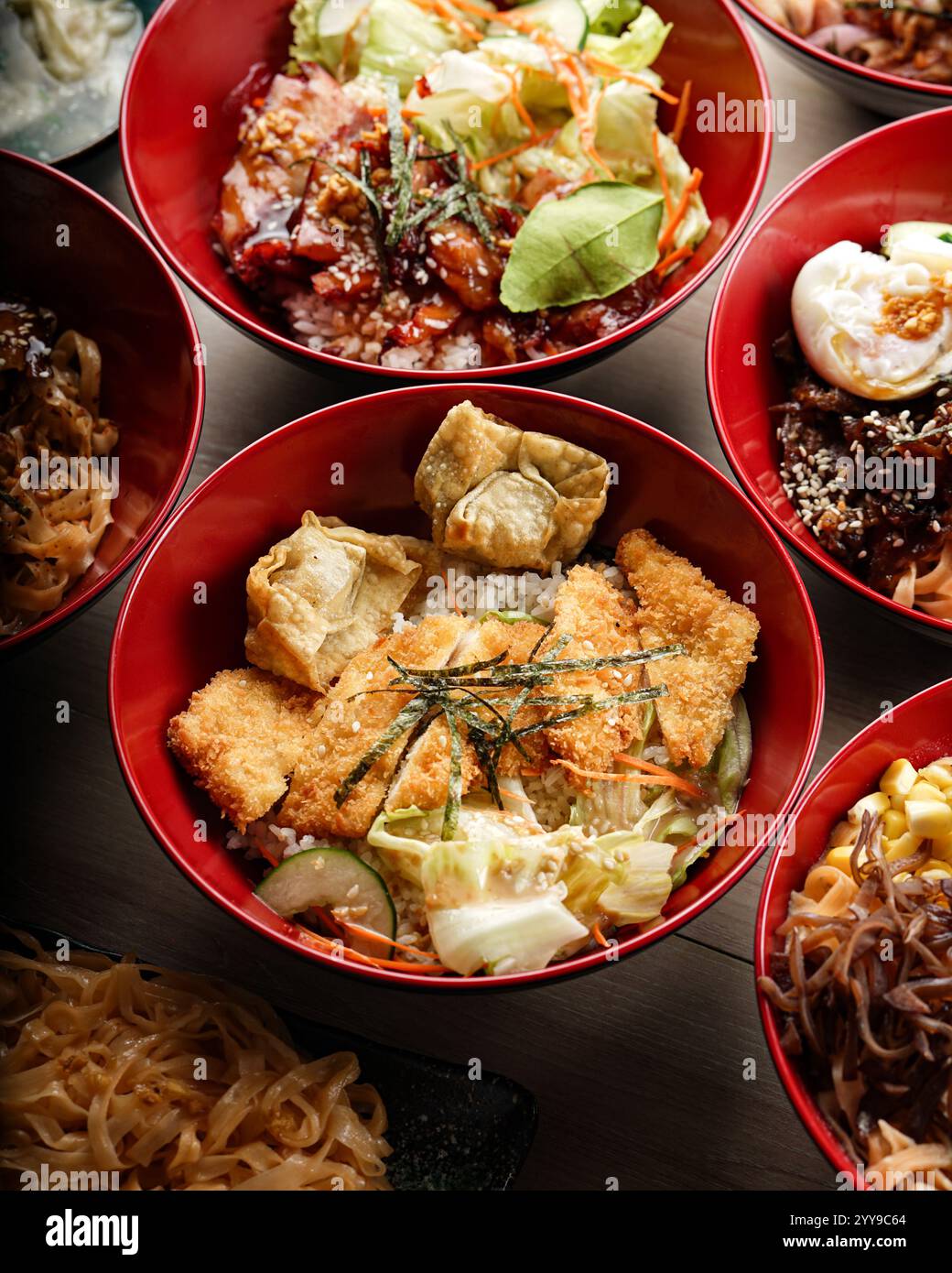 Blick von oben auf eine lebendige asiatische Mahlzeit mit mehreren roten Schüsseln gefüllt mit verschiedenen Reis- und Nudelgerichten, einschließlich gegrilltem Fleisch und Gemüse Stockfoto