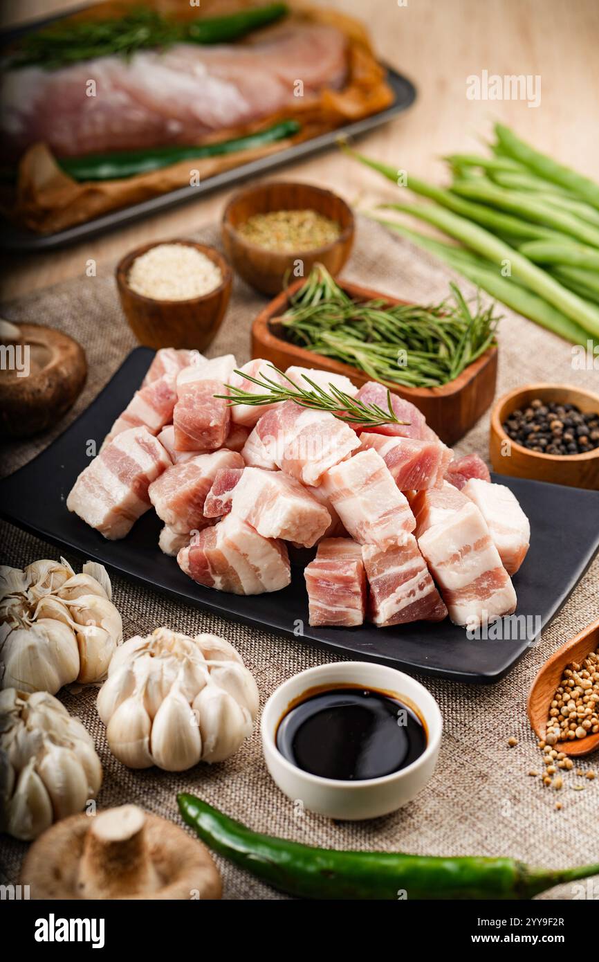 Frisch geschnittene Schweineschulter auf einem rustikalen Holzbrett, umgeben von lebendigem Gemüse und Gewürzen – bereit für eine herzhafte hausgemachte Mahlzeit. Stockfoto