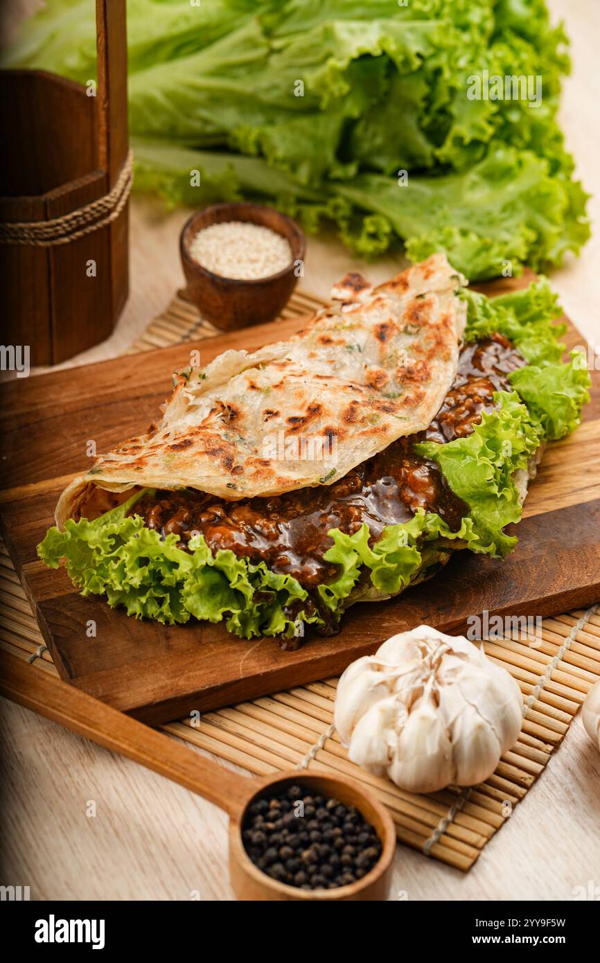 Herzhaftes Rindfleisch, gewickelt in ein blättriges, goldbraunes Fladenbrot mit knusprigem Salat, umgeben von frischen Zutaten wie Knoblauch, Schalotten und Chili Stockfoto