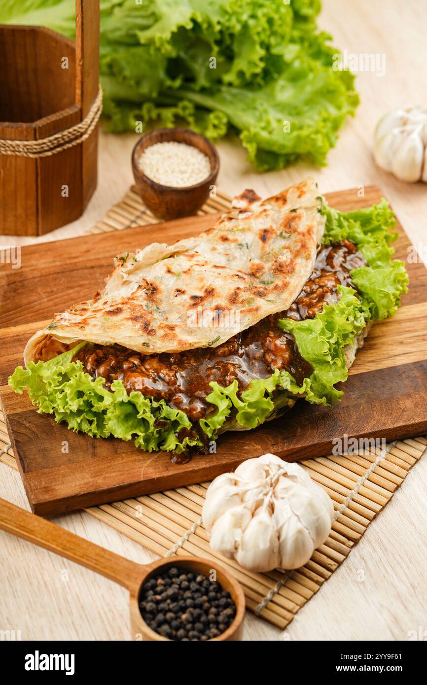 Herzhaftes Rindfleisch, gewickelt in ein blättriges, goldbraunes Fladenbrot mit knusprigem Salat, umgeben von frischen Zutaten wie Knoblauch, Schalotten und Chili Stockfoto