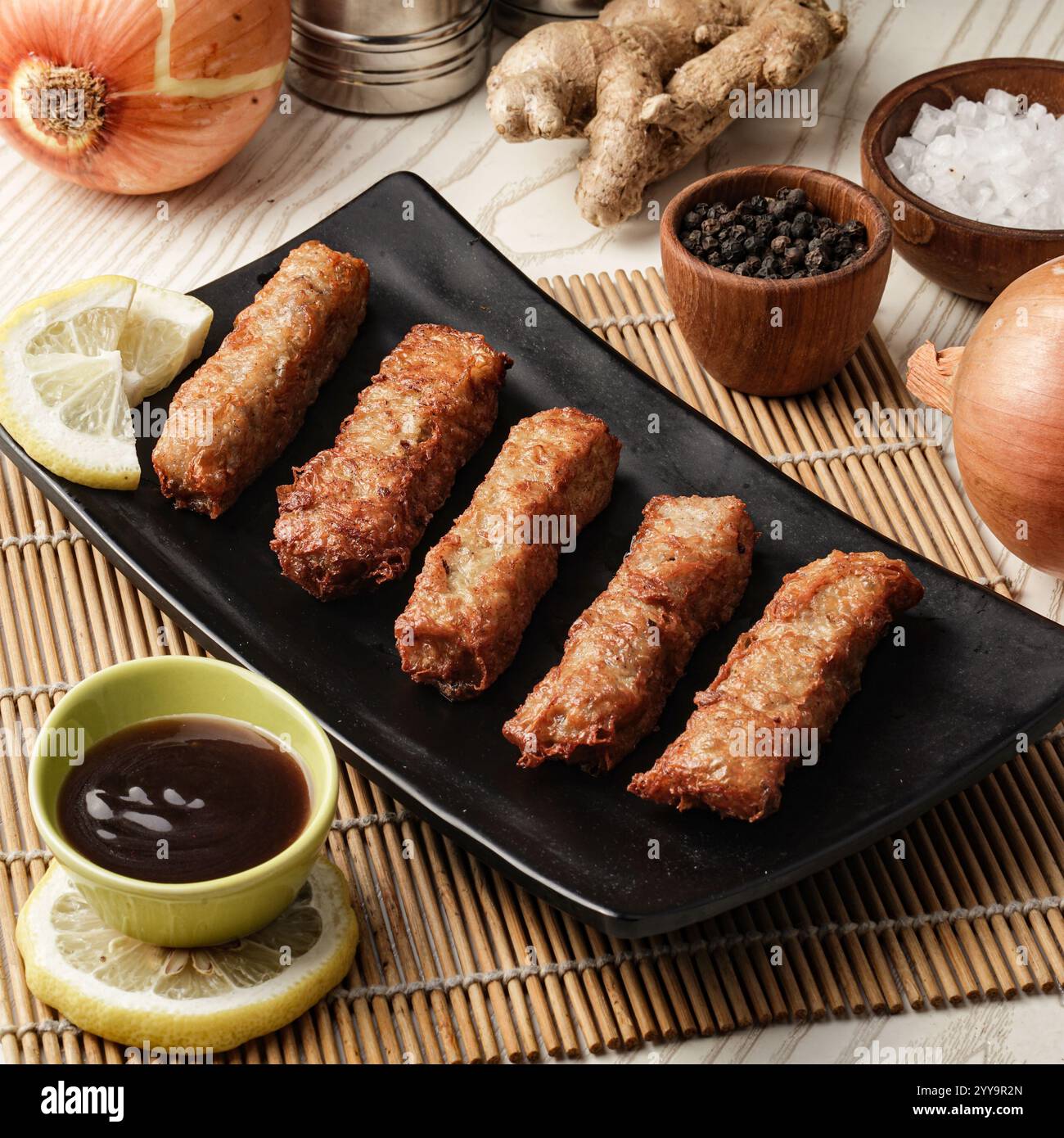 Köstliche knusprige Frühlingsbrötchen serviert auf einem schwarzen Teller mit Sojasauce, garniert mit Zitronenscheiben und umgeben von frischen Zutaten Stockfoto