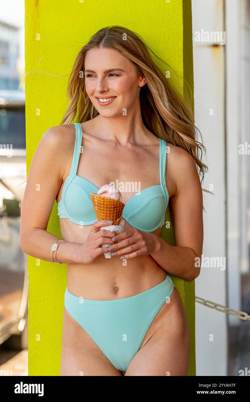 Eine wunderschöne Frau liegt an einem Sandstrand, ihr Bikini betont ihre goldene Bräune. Sie genießt eine Eiskugel, die Wärme der Sonne umschließt sie Stockfoto