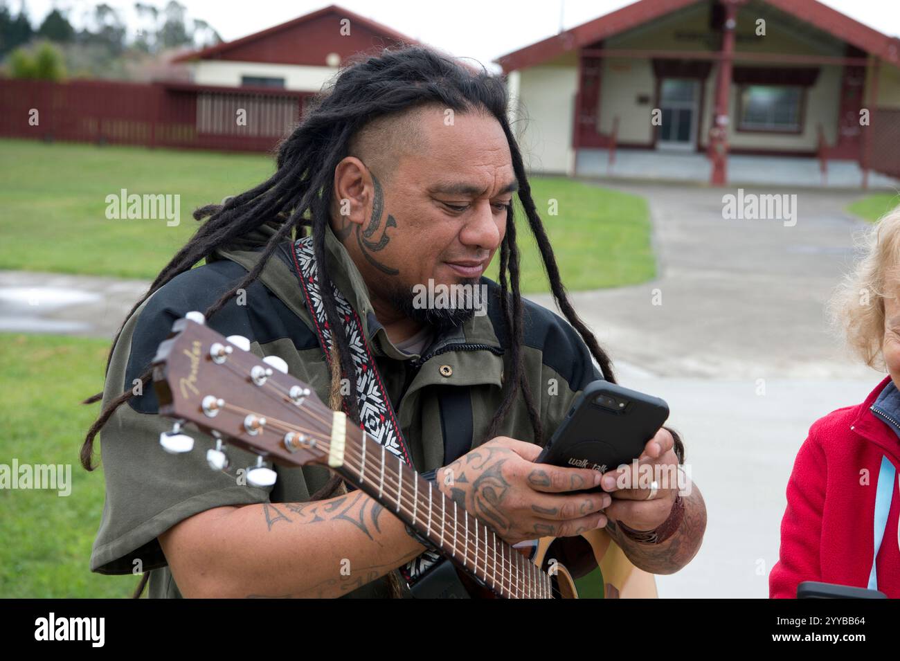 Maori-Mann mit Gitarre, der Handy benutzt, neben einer Frau mit roter Jacke. Stockfoto
