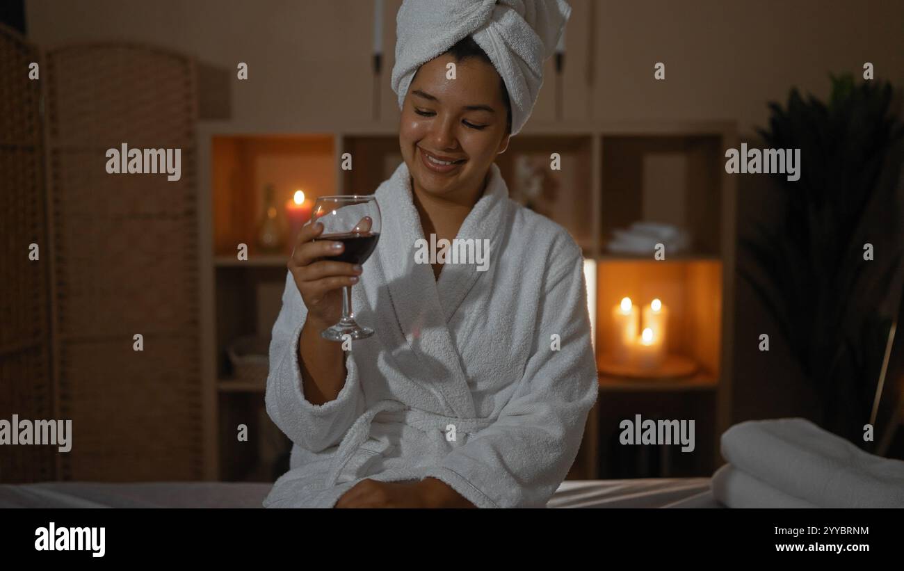 Entspannte hispanische Frau in einem Spa, die einen Bademantel und ein Handtuch auf dem Kopf trägt und ein Glas Rotwein mit Kerzen im Hintergrund genießt. Stockfoto