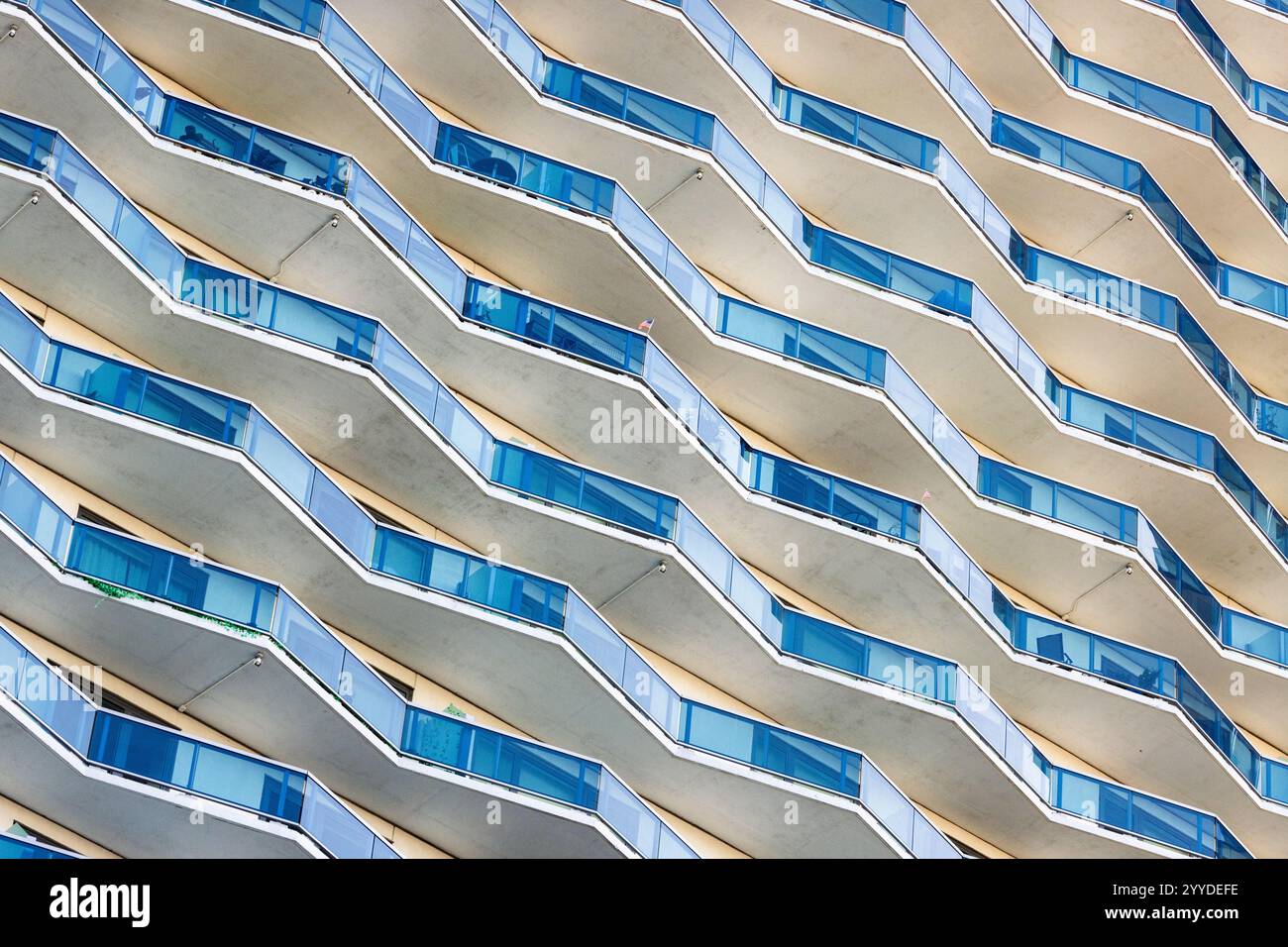 Balkonreihen mit blauen Glasgeländern erzeugen ein sich wiederholendes geometrisches Muster an der Fassade des Gebäudes. Sonnenlicht wirft Schatten und betont das Wa Stockfoto