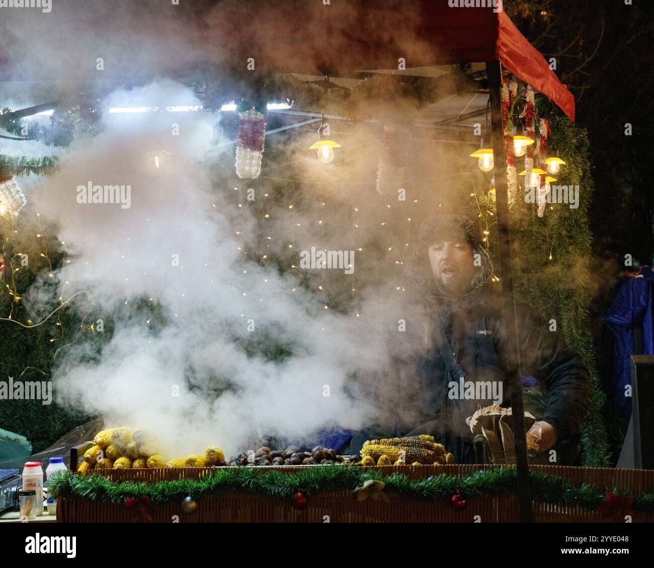Ein festlicher Verkäufer auf Sofias Weihnachtsmarkt, der in Dampf gehüllt und von leuchtenden Lichtern umgeben ist und gerösteten Mais in einer warmen Winterkulisse präsentiert. Stockfoto