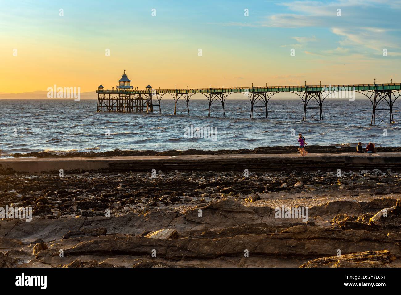 Clevedon Pier in der Goldenen Stunde bei Sonnenuntergang mit sonnendurchflutetem Ufer und Menschen, die den Bootssteg hinunterlaufen Stockfoto