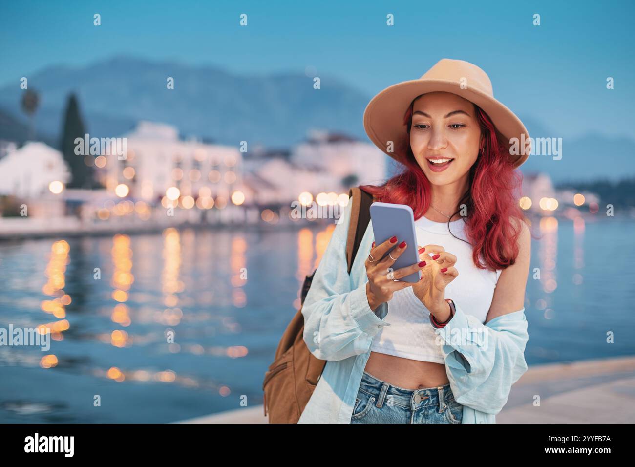 Junge Frau, die in der Abenddämmerung vor den Lichtern am Meer ein Smartphone benutzt Stockfoto