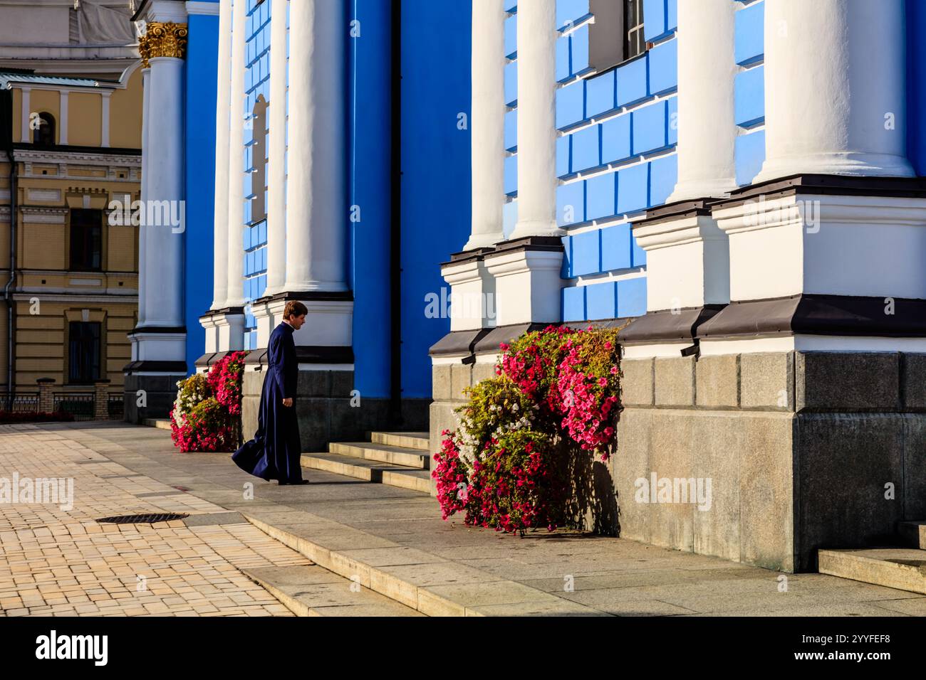 Eine Frau in langem Kleid geht an einem Gebäude mit einer blau-weißen Fassade vorbei. Das Gebäude hat ein großes Fenster auf der Vorderseite und einen Balkon mit Topf pla Stockfoto