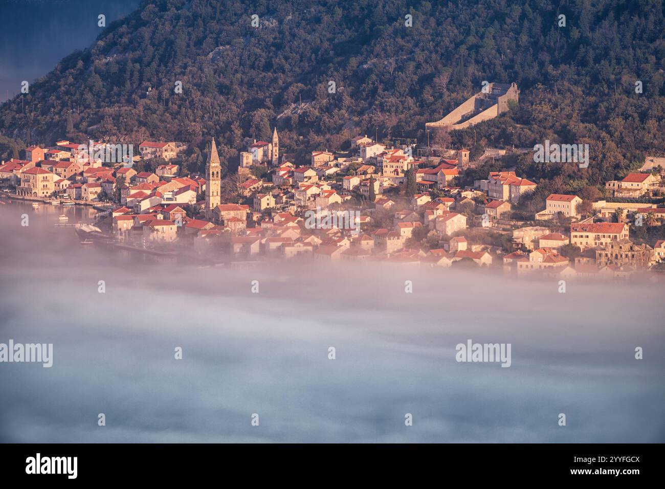 Malerischer Blick auf das Dorf Perast, das bei Sonnenaufgang teilweise von Nebel bedeckt ist, mit seinen charakteristischen roten Dächern und Kirchtürmen in der Bucht von Kot Stockfoto