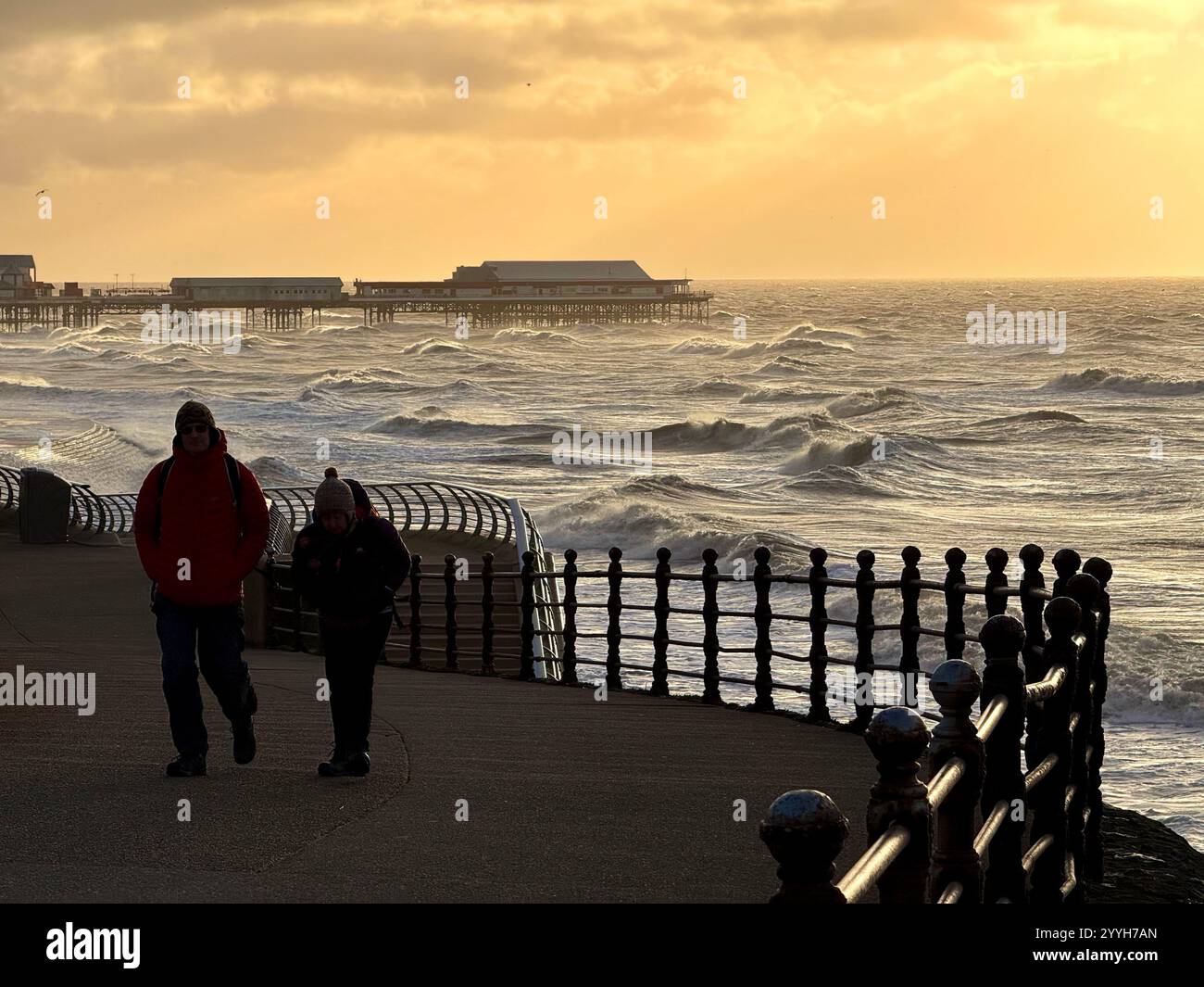 Zwei Personen laufen bei Flut an einem windigen Winterabend auf der Blackpool Promenade mit North Pier in der Ferne Stockfoto