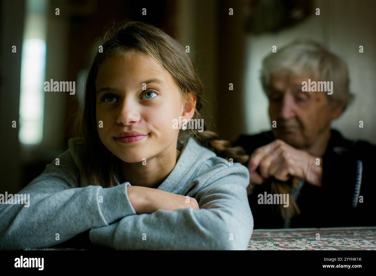 Eine Großmutter flechtet die Haare ihrer Enkelin, während der Teenager mit einem etwas unzufriedenen Ausdruck sitzt, der in einem ruhigen, intimen Moment festgehalten wird. Stockfoto