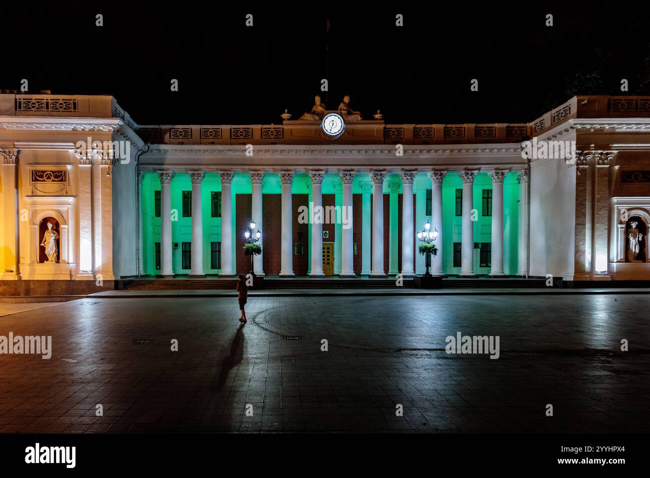 Ein großes Gebäude mit grüner Fassade wird nachts beleuchtet. Eine Person läuft vor dem Gebäude Stockfoto