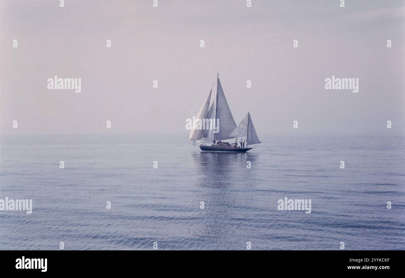 Historisches Foto des ruhigen Meerwassers mit einem Segelboot mit zwei Masten, aufgenommen in den 1960er Jahren in England, Großbritannien Stockfoto