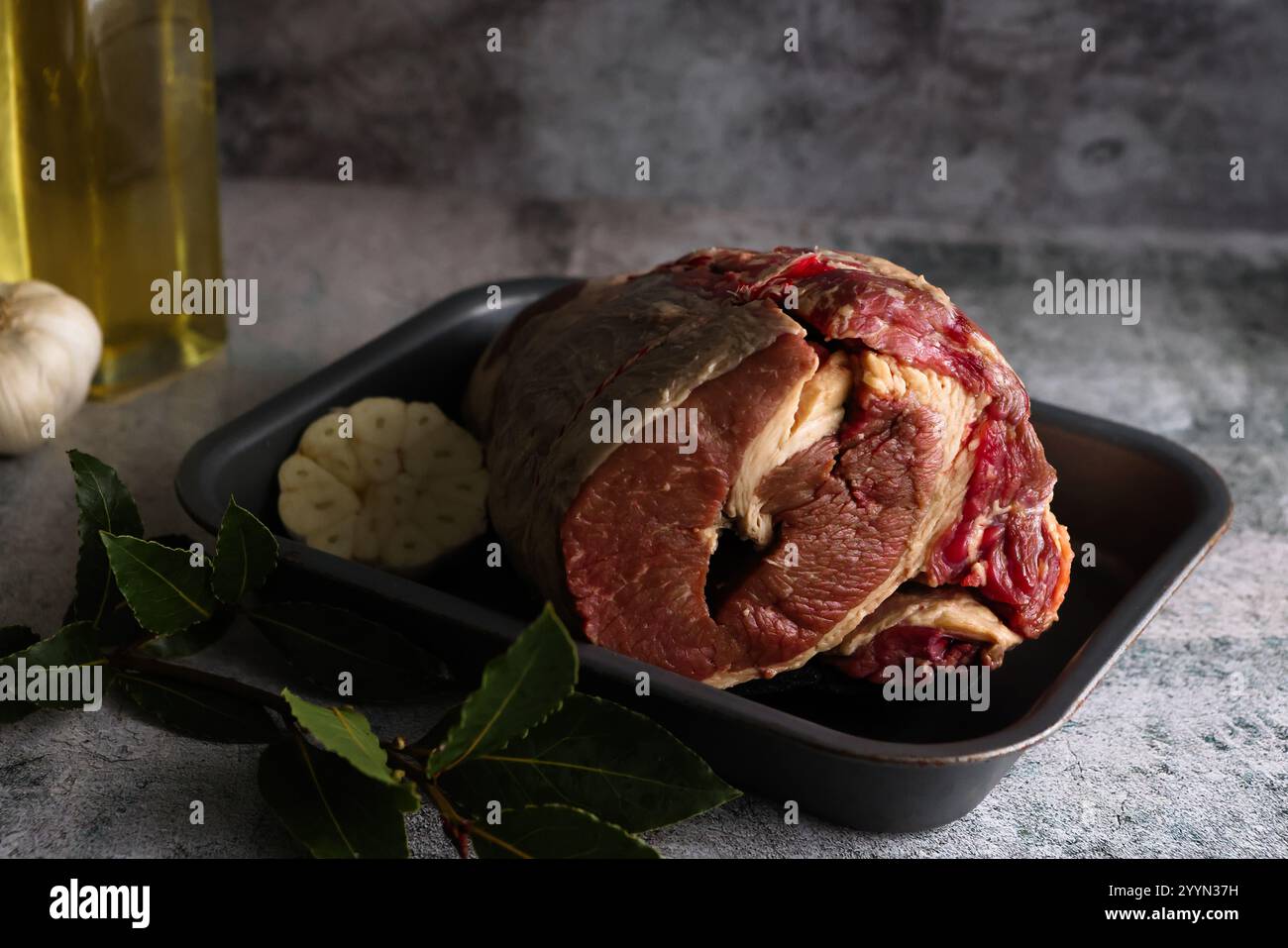 Sonntags Rinderbraten in einer Straßenform, roh und mit einer ganzen Galliskolbe und Kräutern auf grauem Hintergrund mit Gewürzen. Stockfoto