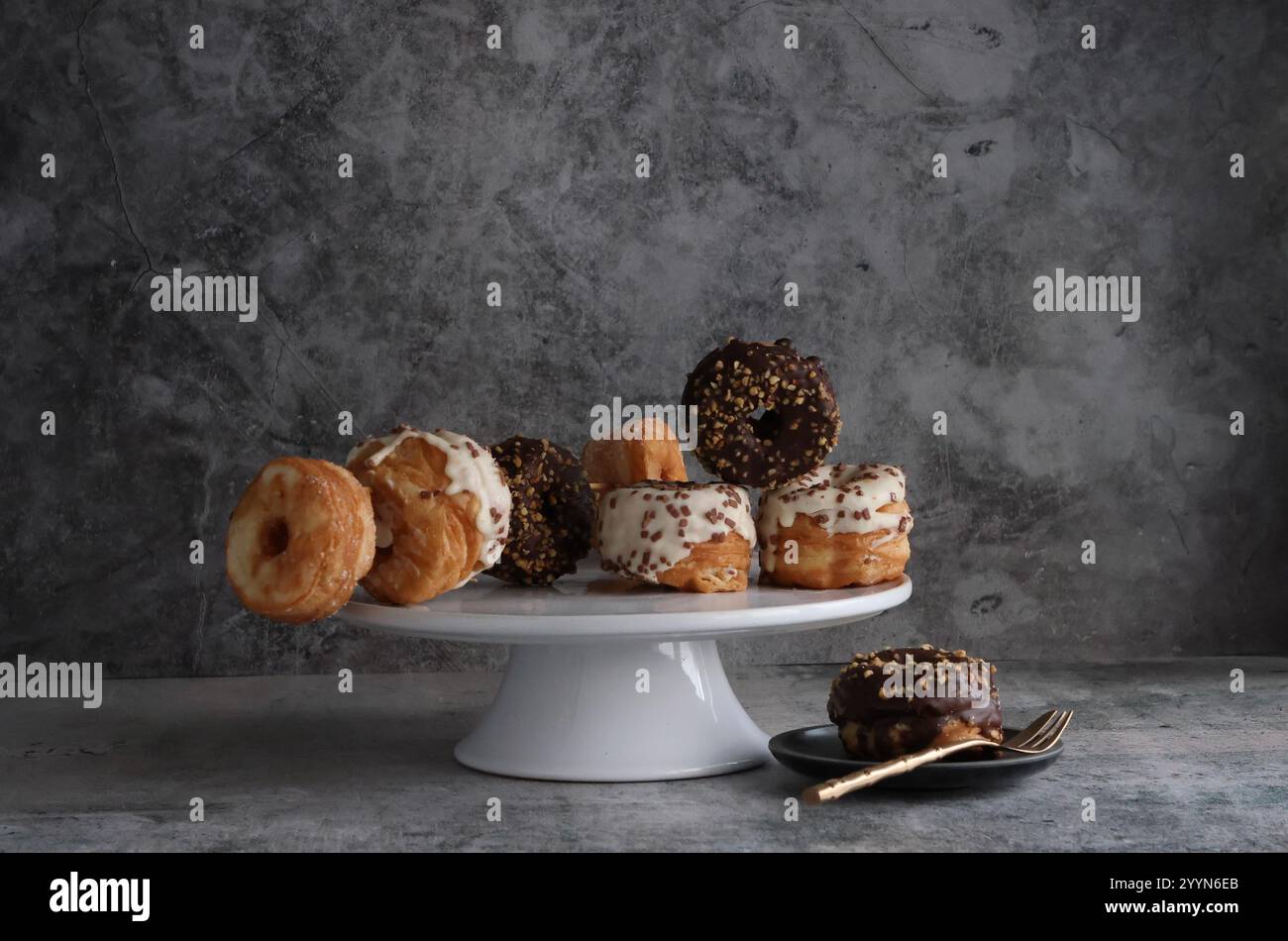 Eine Auswahl an Crodots oder Mini Donuts auf einem Kuchenständer mit verschiedenen Glasur und Streuseln. Eine Beilage mit vergoldeter Gabel und einem Kuchen. Stockfoto