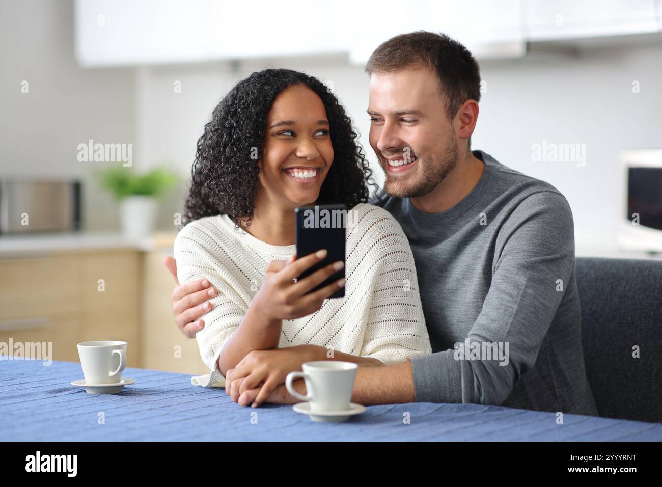 Glückliches interrassisches Paar, das telefoniert und sich zu Hause in der Küche ansieht Stockfoto