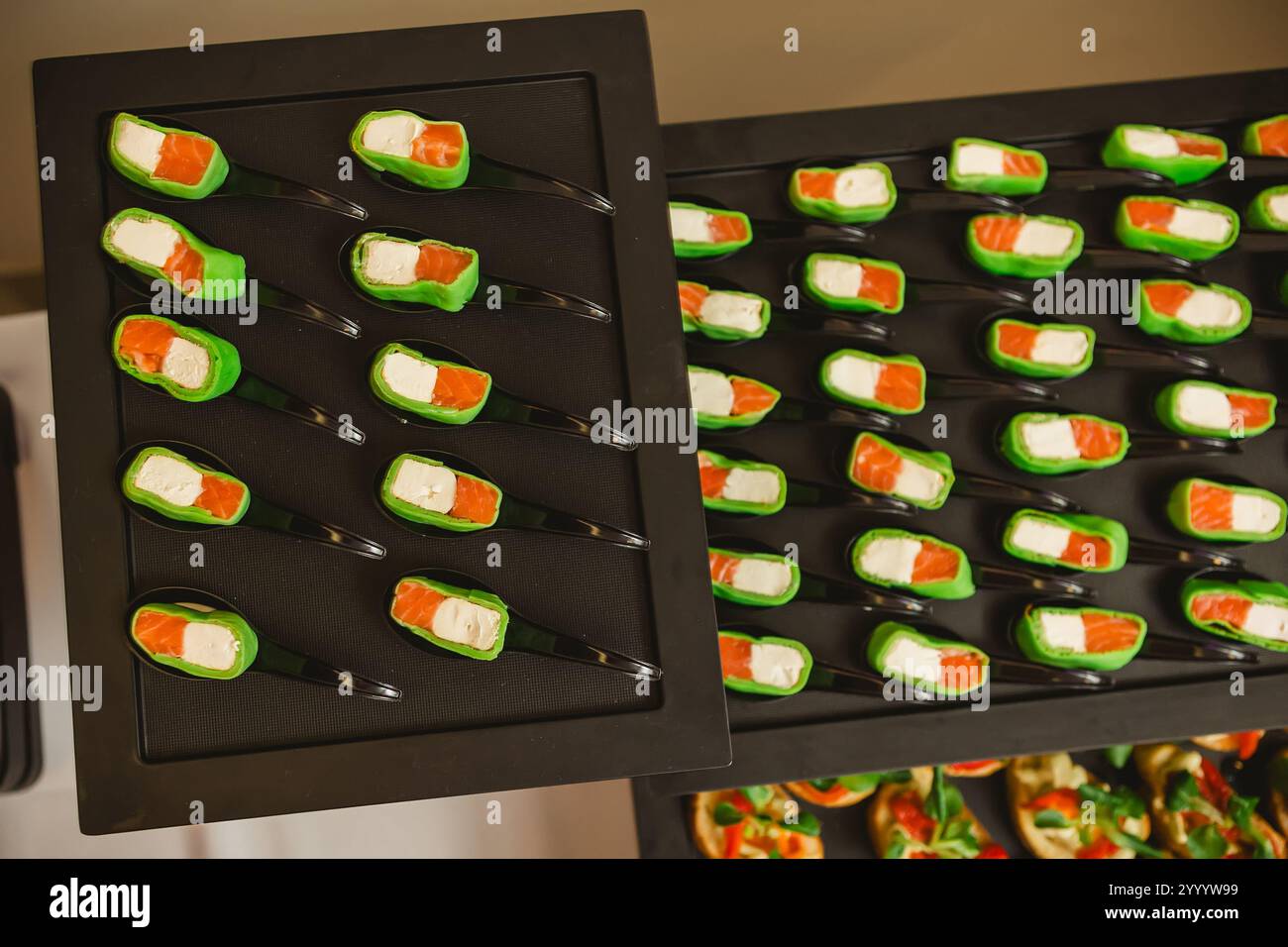 Platte mit Gourmet-Vorspeisen mit Lachsstücken und Frischkäse in einer grünen Verpackung, serviert auf schwarzen Löffeln. Die Präsentation ist elegant Stockfoto