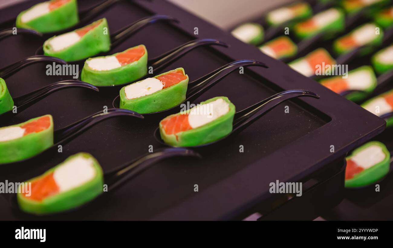 Platte mit Gourmet-Vorspeisen mit Lachsstücken und Frischkäse in einer grünen Verpackung, serviert auf schwarzen Löffeln. Die Präsentation ist elegant Stockfoto
