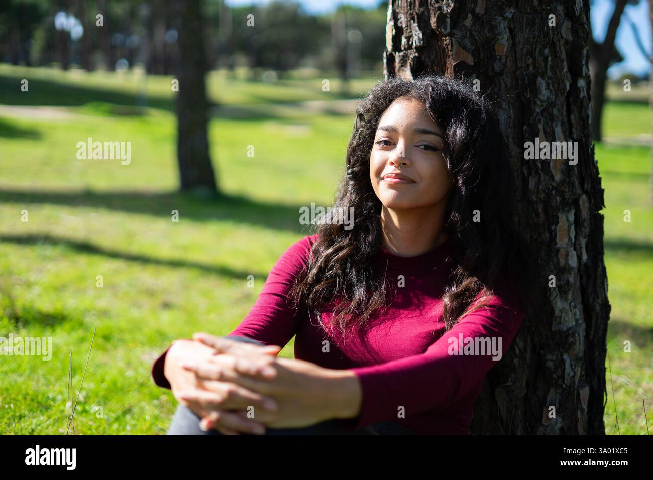 Eine ruhige junge lateinfrau genießt einen friedlichen Moment in der Natur, sitzt auf Gras und lehnt sich an einen Baum im Park Stockfoto