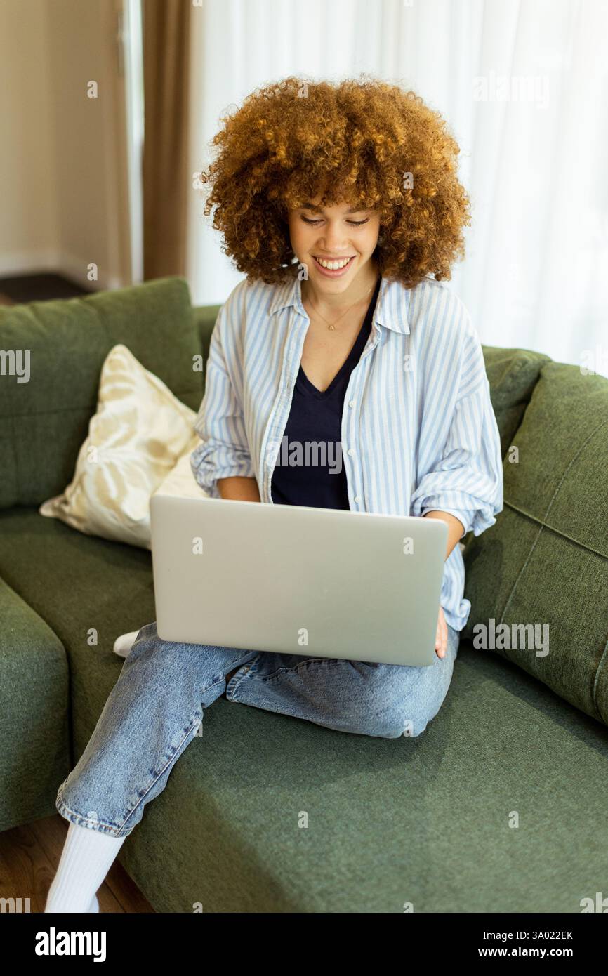 Eine junge Frau sitzt bequem auf einem weichen, grünen Sofa und tanzt sich auf ihrem Laptop, sonnt sich im warmen Sonnenlicht, das durch die großen Fenster strömt Stockfoto